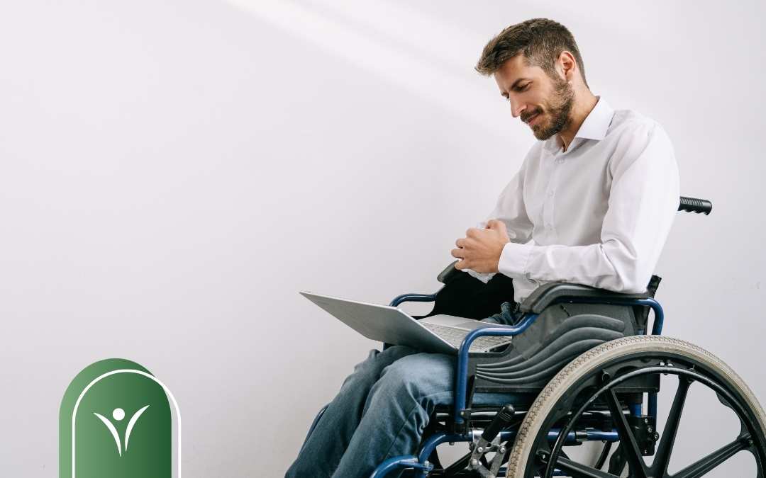 Mężczyzna na wózku inwalidzkim pracujący na laptopie, siedzący przy ścianie w jasnym pomieszczeniu.