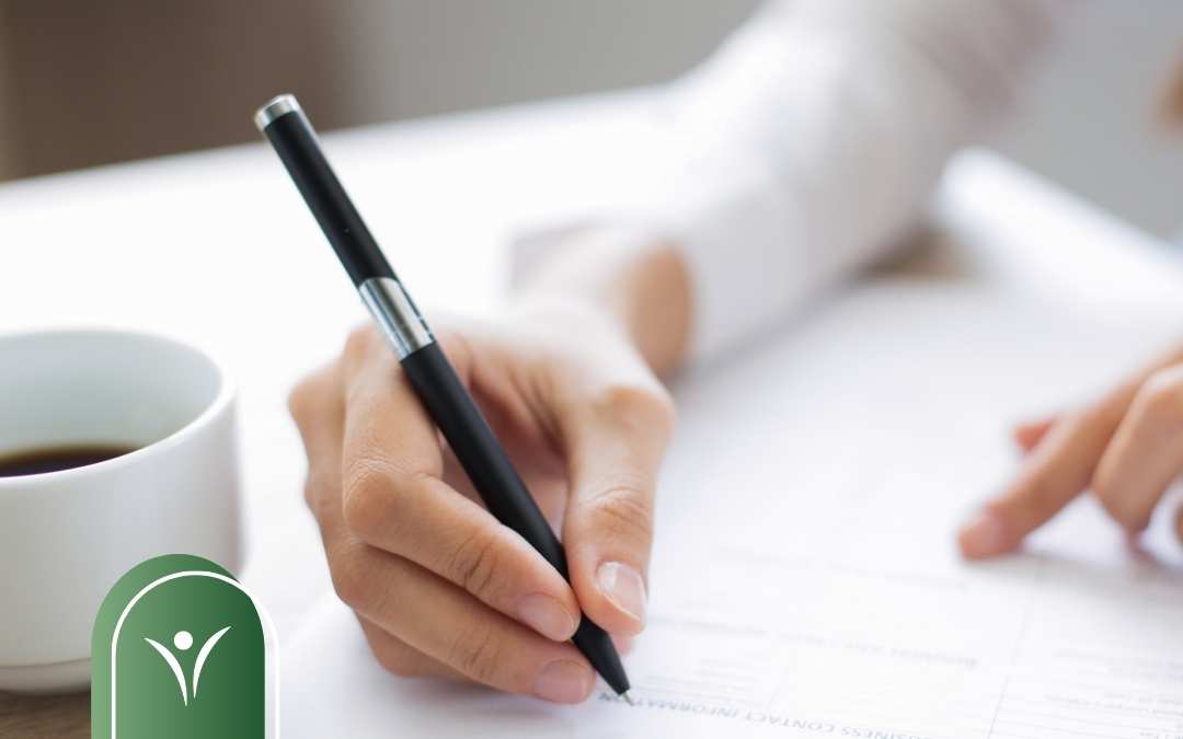 Dłoń osoby dorosłej wypełniającej dokument papierowy długopisem, na biurku widoczna filiżanka kawy; grafika ilustruje składanie wniosku o świadczenie wspierające.