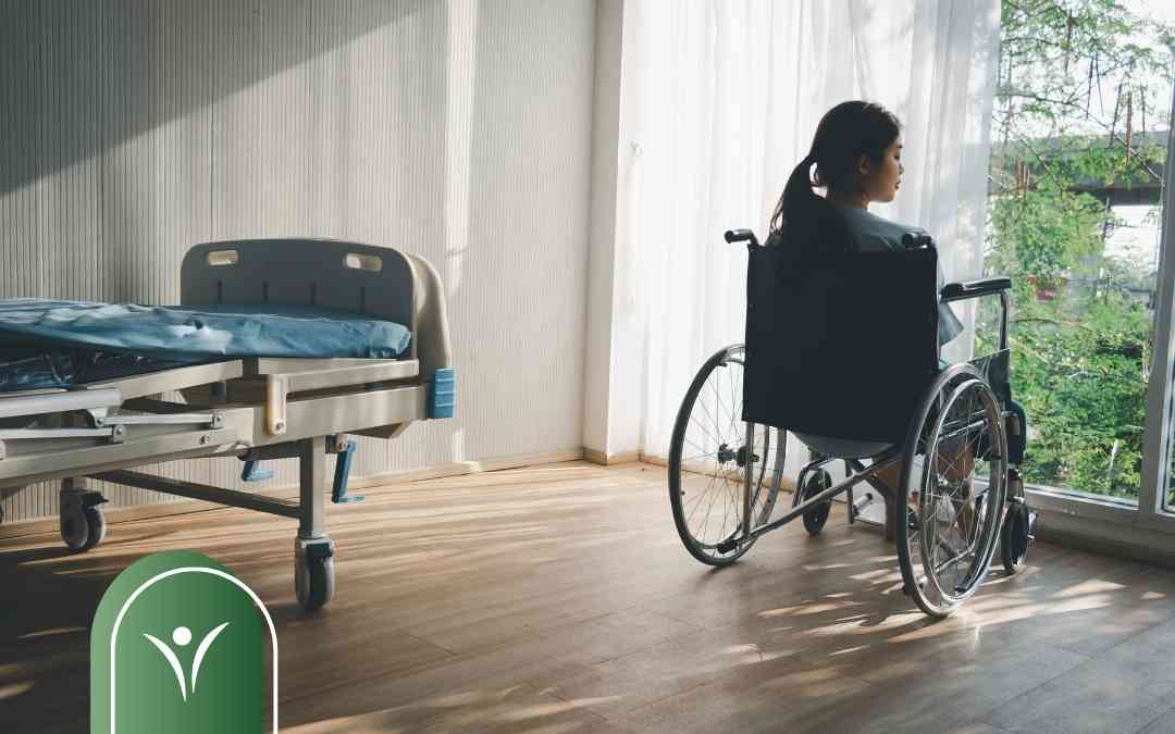Kobieta na wózku inwalidzkim patrzy przez okno w pokoju z łóżkiem rehabilitacyjnym.
