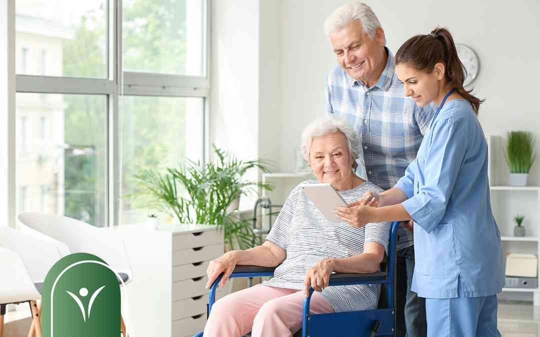 Nurse in blue scrubs shows a tablet to an elderly woman in a wheelchair while a man smiles behind them in a bright room.