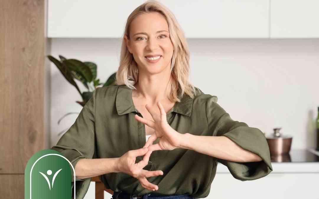 Uśmiechnięta kobieta pokazująca znak w języku migowym na tle jasnej kuchni.