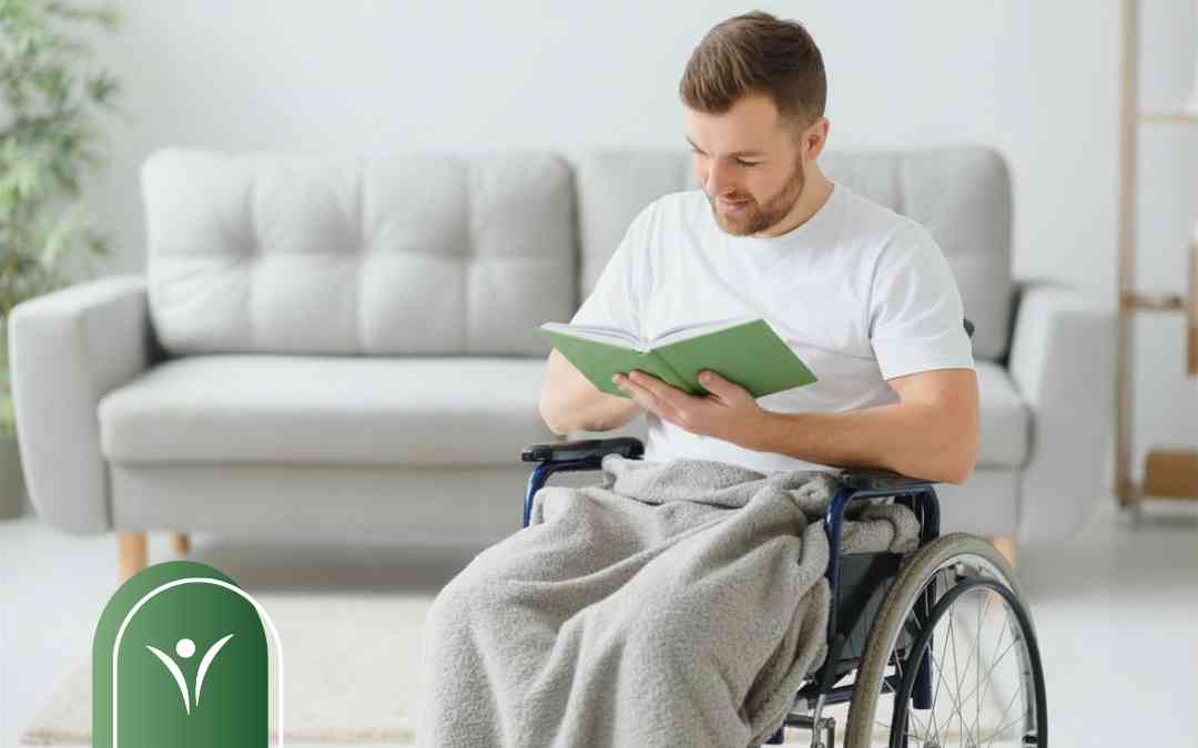 Mężczyzna na wózku inwalidzkim czyta zieloną książkę, ilustrując samodzielność w codziennych czynnościach.