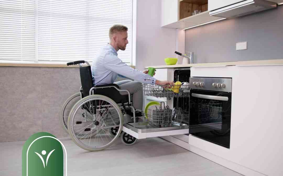 Man in a wheelchair loading a dishwasher in a bright modern kitchen with white cabinets and a stainless oven.