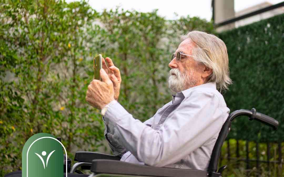 Elderly man with white hair and beard sits in a wheelchair outdoors, holding a smartphone to take a photo in a garden.