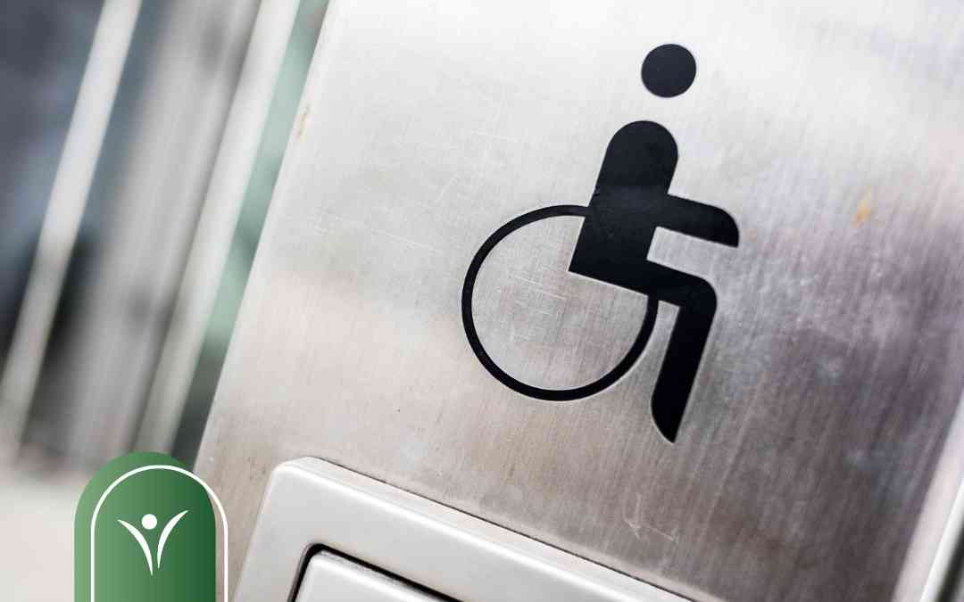 Czarna ikona osoby na wózku inwalidzkim na srebrnej, metalowej tabliczce informacyjnej.