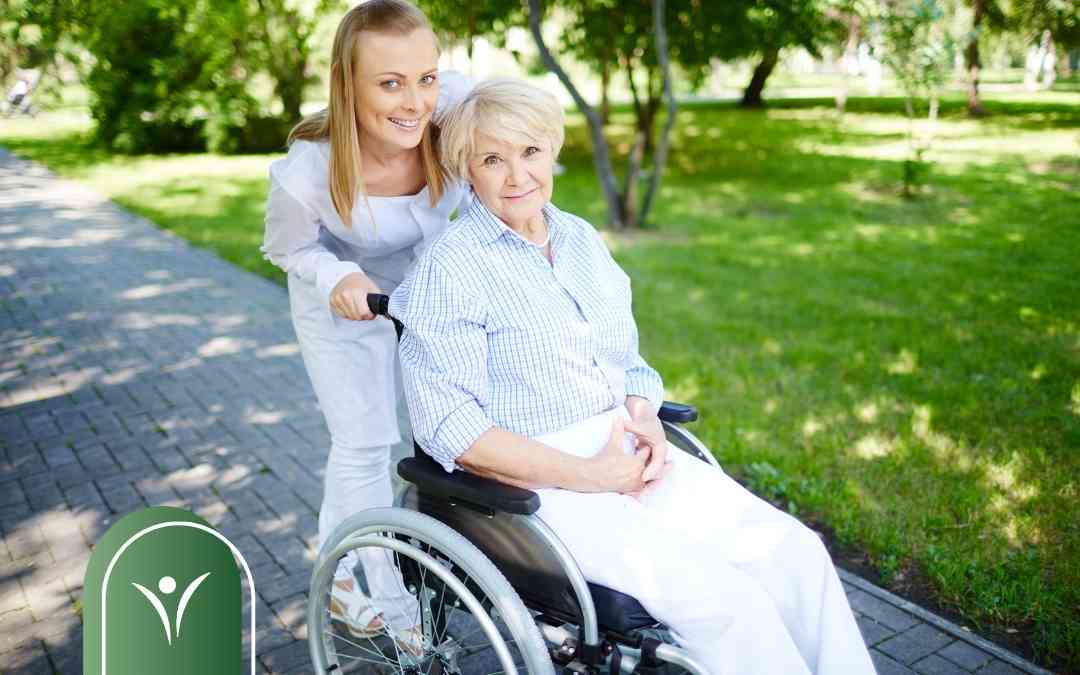 Uśmiechnięta opiekunka prowadzi wózek inwalidzki ze starszą kobietą po parkowej alejce.