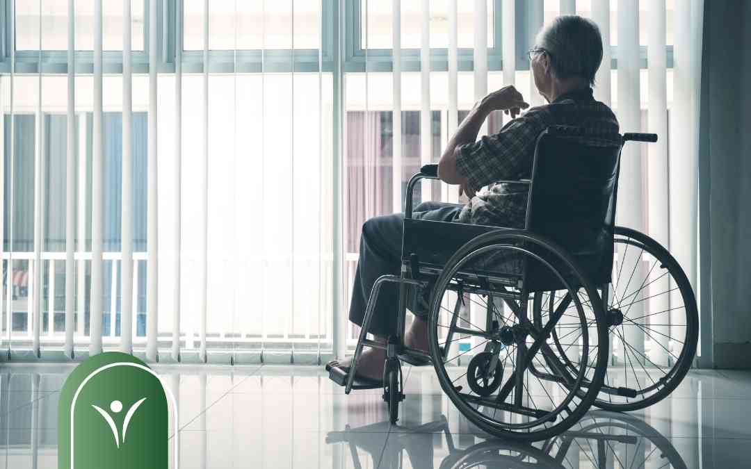 Starszy mężczyzna na wózku inwalidzkim patrzy przez okno z żaluzjami w jasnym korytarzu.