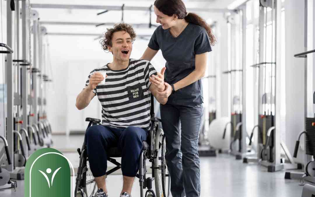 Uśmiechnięty mężczyzna na wózku inwalidzkim ćwiczy z fizjoterapeutką w nowoczesnej sali rehabilitacyjnej.