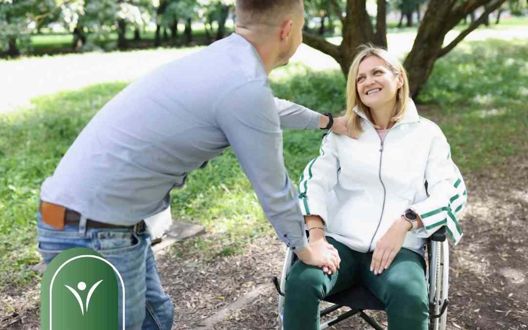 Uśmiechnięty mężczyzna wspiera kobietę na wózku inwalidzkim, trzymając ją za rękę w parku.