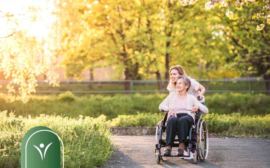 Kobieta spaceruje w słonecznym parku z seniorką na wózku inwalidzkim – wsparcie osoby z niepełnosprawnością