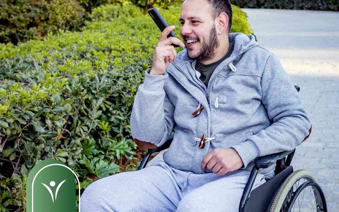 Mężczyzna na wózku inwalidzkim rozmawia przez telefon w trybie głośnomówiącym na tle zieleni.